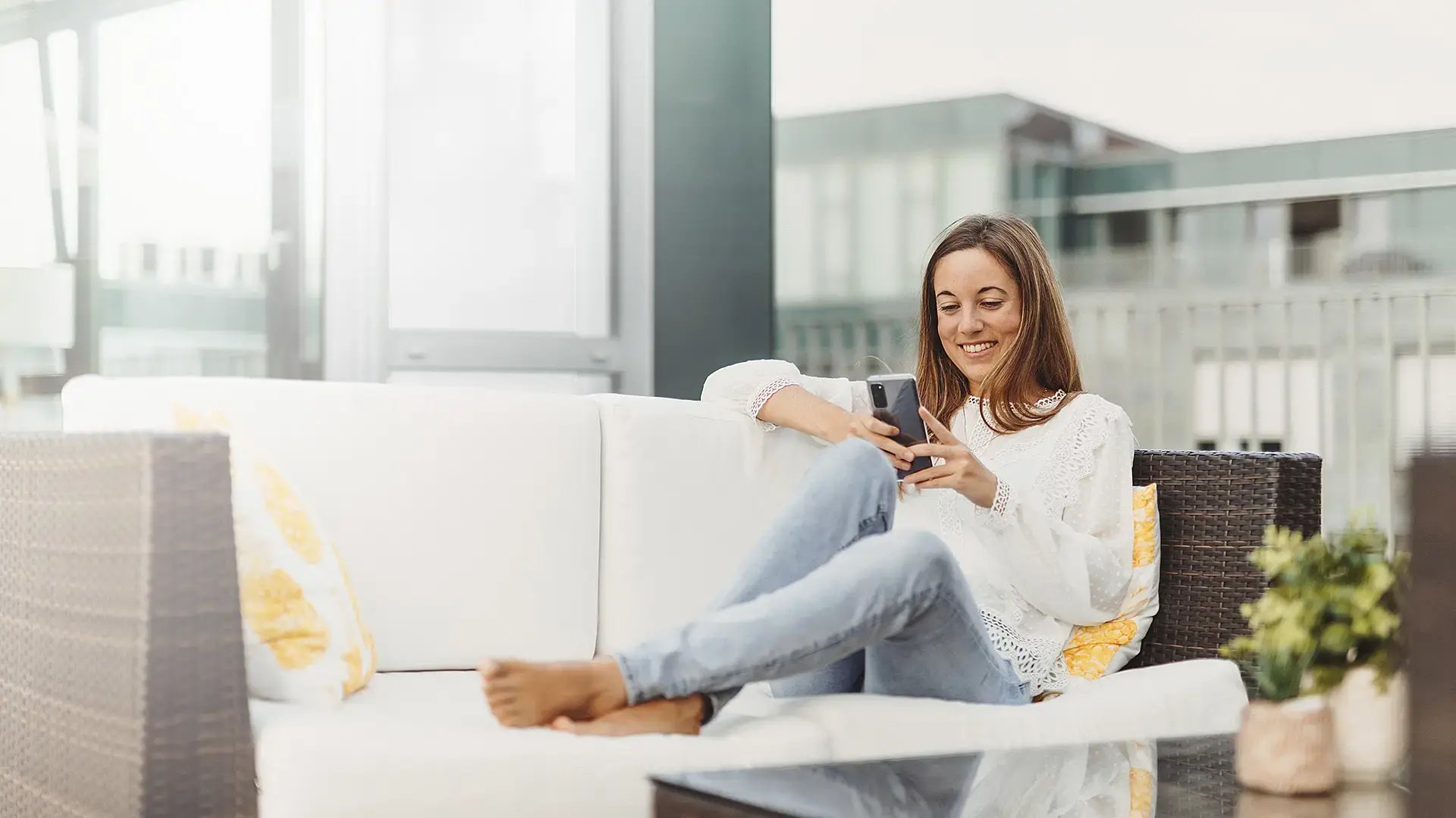 Een vrouw zit op een bank. Ze kijkt op haar mobiel en lacht.