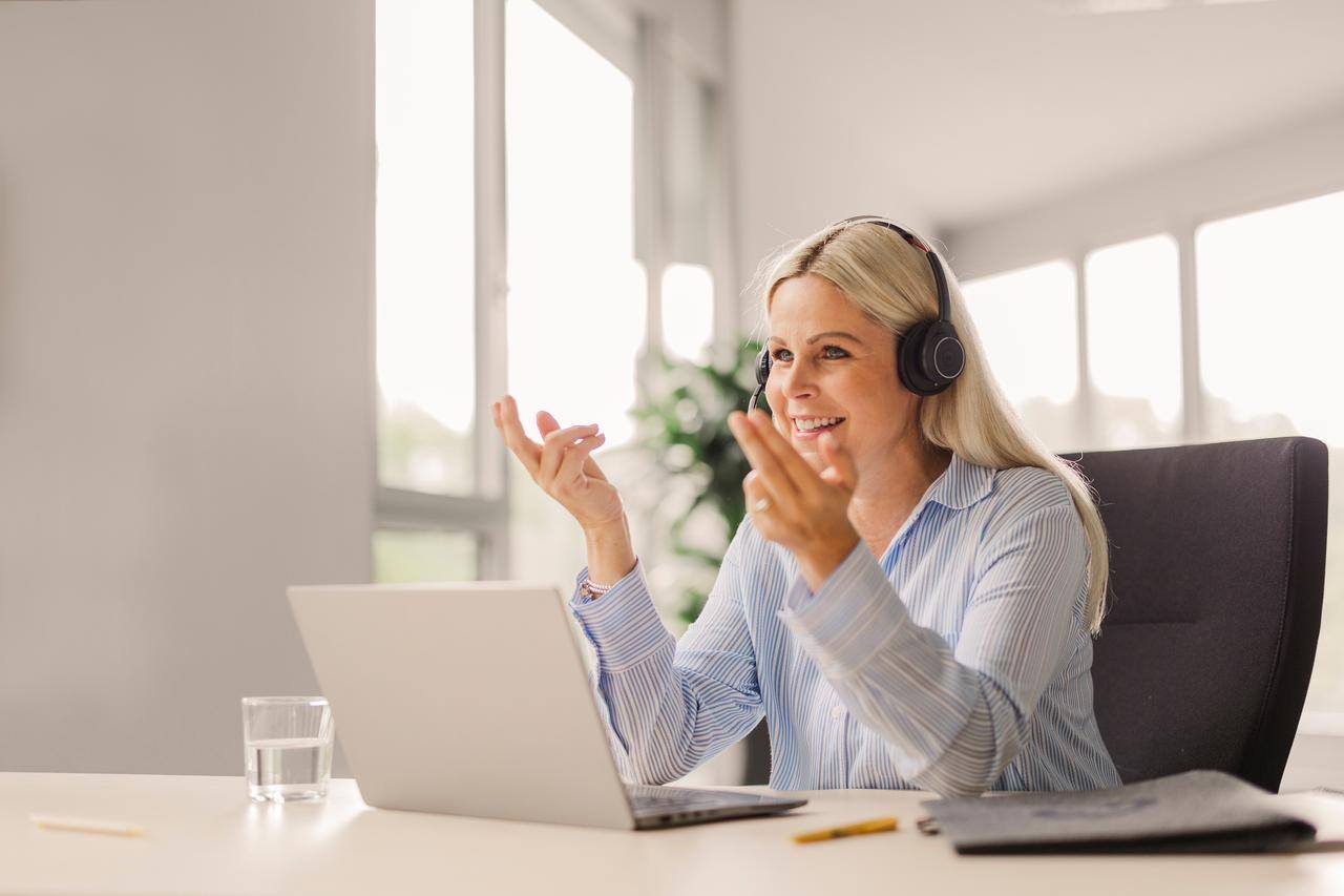 Afbeelding van een vrouw die achter een laptop zit, met een headset op. Ze lacht en lijkt iets uit te leggen.