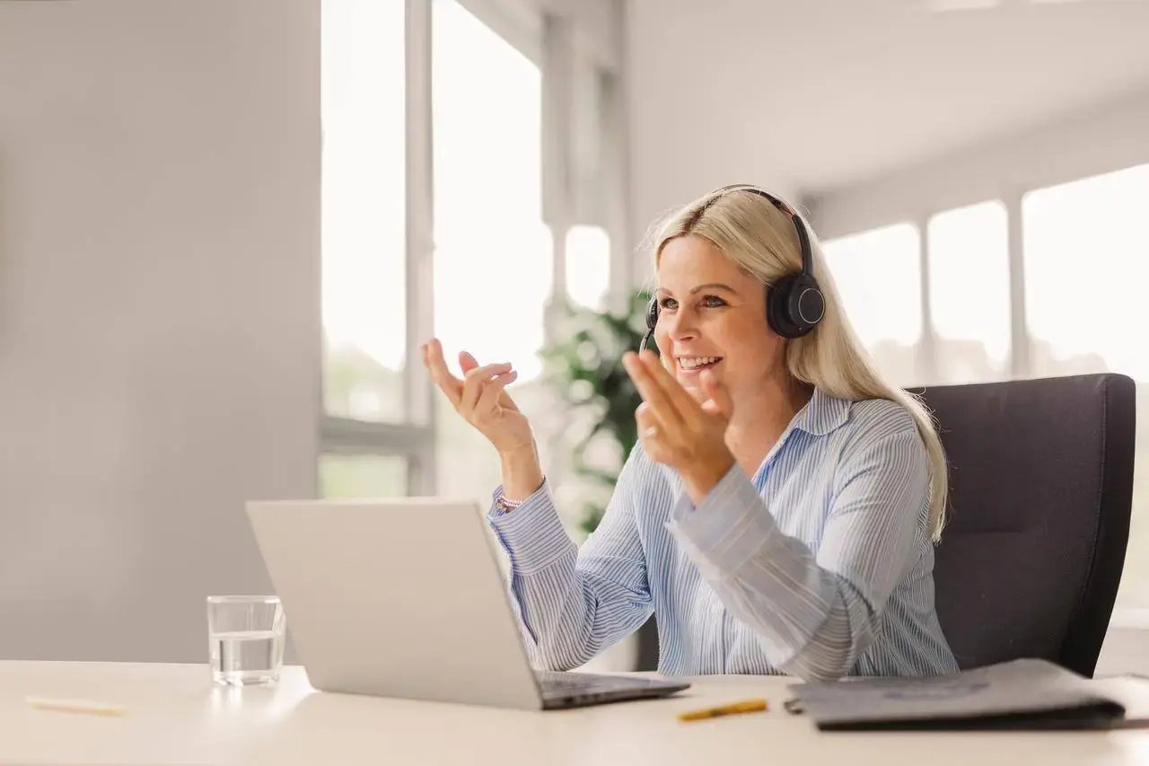 Afbeelding van een vrouw die achter een laptop zit, met een headset op. Ze lacht en lijkt iets uit te leggen.