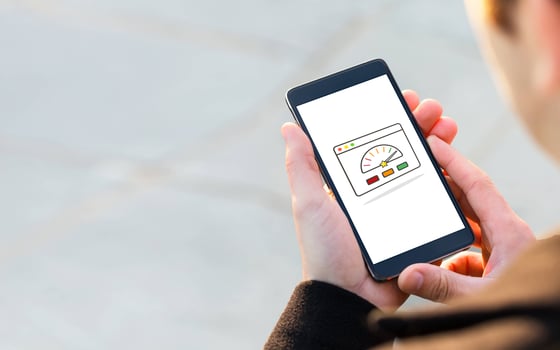Persoon houdt smartphone vast met dashboard en snelheidsmeter-icoon op het scherm, symbool voor websiteverkeer en performance. Beeld visualiseert traffic, groei en online statistieken.