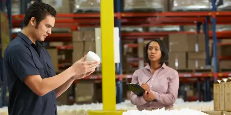 Medewerkers in een magazijn beoordelen een nieuw product en maken aantekeningen tijdens het testen van productvraag voor een webshop. Dozen en stellingen op de achtergrond benadrukken logistiek en voorraadbeheer.