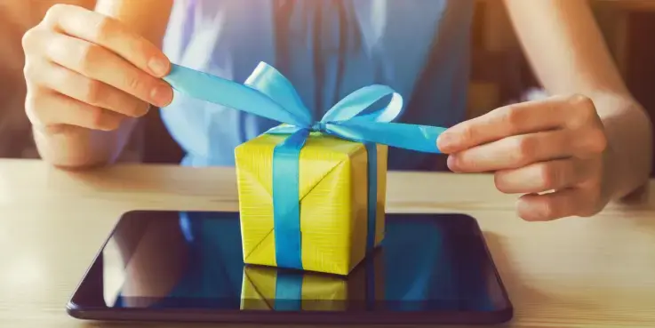 Persoon pakt een klein geel cadeau met blauwe strik op een zwarte tablet; symbolische afbeelding voor loyaliteitsprogramma’s, klantenbinding en beloningssystemen voor webshops.