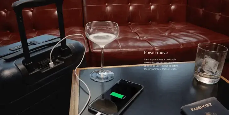 Afbeelding van Away Travel met een handbagagekoffer met ingebouwde USB-oplader die een smartphone oplaadt, op een tafeltje met een paspoort en glazen drankjes in een lounge-achtige setting. Benadrukt slim design en reizen met power.