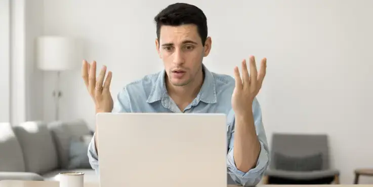Een man zit achter een laptop, met een lichtblauwe bloes met lange mouwen, hij heeft kort donker haar. Hij heeft z'n handen omhoog in verwarring. Hij weet niet hoe hij reageert op negatieve beoordelingen.