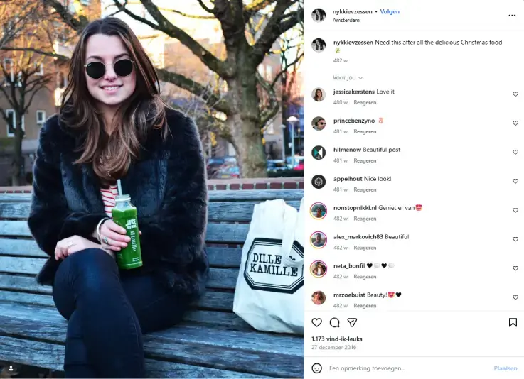 Instagramfoto van fake influencer Nykkievzessen die op een houten bank in Amsterdam zit met een groene juice in de hand en een Dille & Kamille-tas naast zich, met winterse bomen en stedelijke omgeving op de achtergrond.