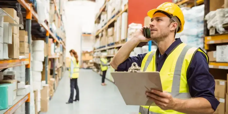 Medewerker in een magazijn controleert voorraden met een clipboard en telefoon, omringd door hoge stellingen en collega’s die producten verwerken. Logistiek proces in een moderne opslagruimte.