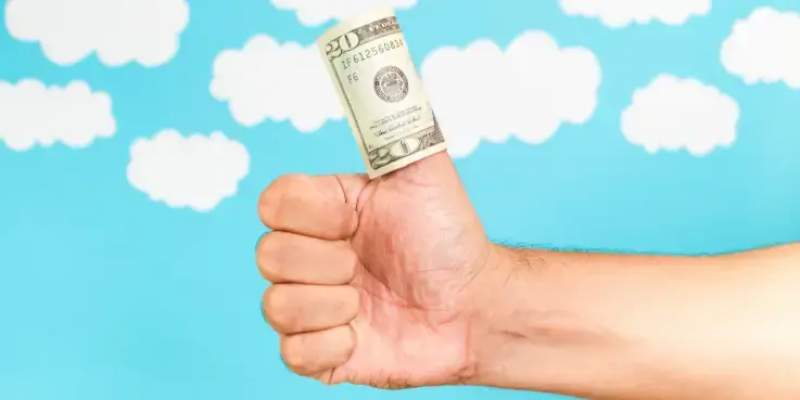 Hand die een opgeheven duim maakt met een opgerolde dollarbiljet eromheen, tegen een blauwe lucht met wolken; symbool voor het bepalen en optimaliseren van een marketingbudget.