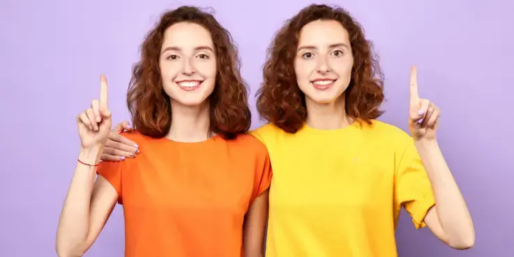 Foto van een tweeling als symbool voor canonical tags. Links heeft een oranje t-shirt, rechts een geel t-shirt. Beide vrouwen hebben halflang golvend rood haar. Ze glimlachen en steken een vinger op.