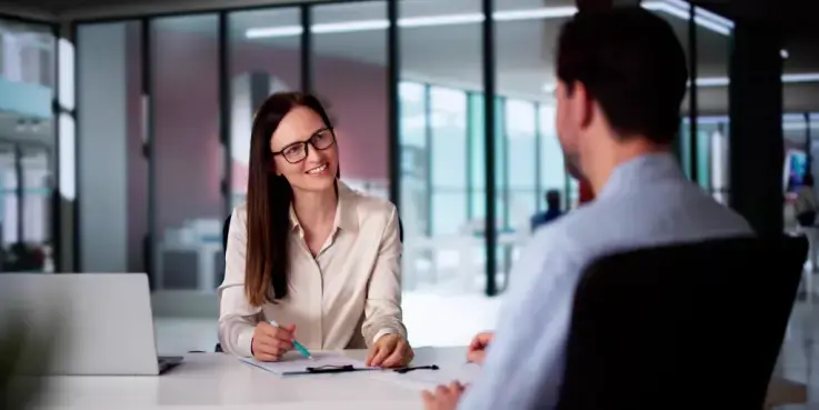 Een vrouw met lang bruin haar en een zwarte bril op houdt een sollicitatiegesprek met een man die we geblurd zien, met de rug naar de camera.
