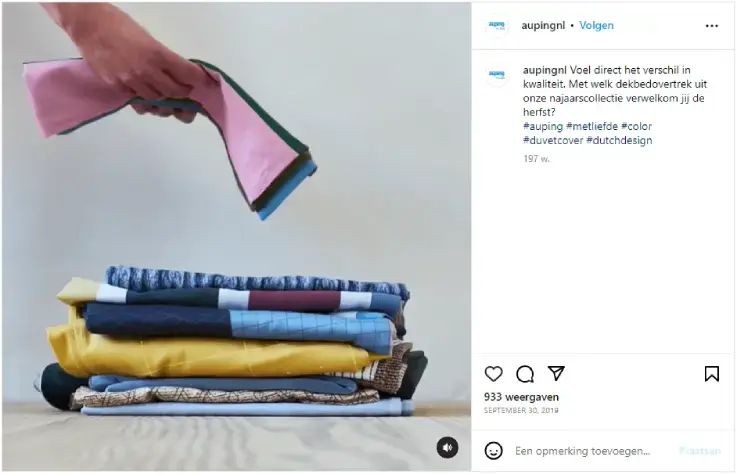 Instagram-post van het account aupingnl met een stapel kleurrijke dekbedovertrekken op de vloer en een hand die een opgevouwen overtrek erboven houdt. Minimalistische setting met zachte kleuren, passend bij Auping najaarscollectie, kwaliteit, beddengoed en herfstinterieur.