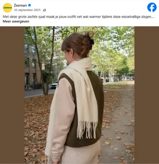 Persoon loopt in een park met herfstbladeren en draagt een lichte jas, groene bodywarmer en een zachte crèmekleurige sjaal van Zeeman. Op de achtergrond staan bomen en gebouwen langs een pad, passend bij een frisse herfstdag.