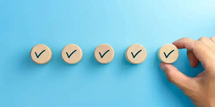 Hand plaatst houten schijfje met vinkje naast vier andere vinkjes op een blauwe achtergrond, als symbool voor het behalen van een webshopkeurmerk en het voldoen aan belangrijke kwaliteits- en veiligheidscriteria voor webshops.
