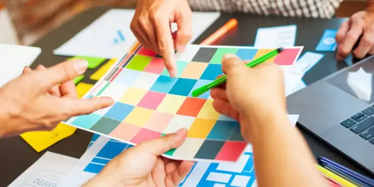 Team dat samen een kleurrijk designpalet bespreekt tijdens een ontwerpsessie, met kleurstalen, schetsen en laptop op tafel. Creatief proces voor webdesign en branding.