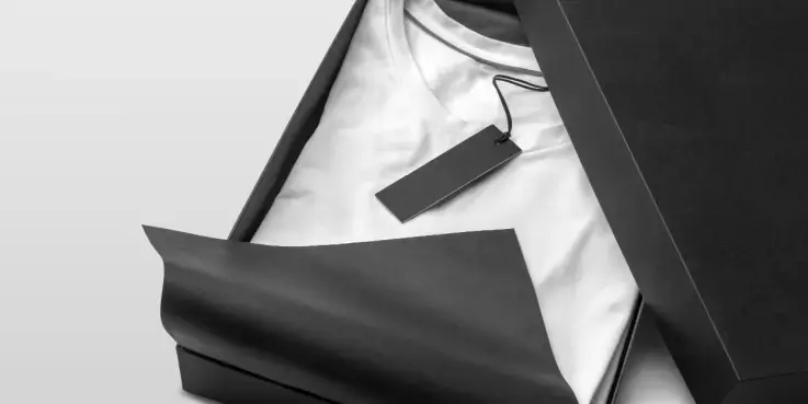 Zwart luxe doosje met daarin een wit T‑shirt, zorgvuldig verpakt in zwart vloeipapier, met een minimalistisch merklabel. Premium productpresentatie voor sterk merk‑ en verpakkingsdesign.